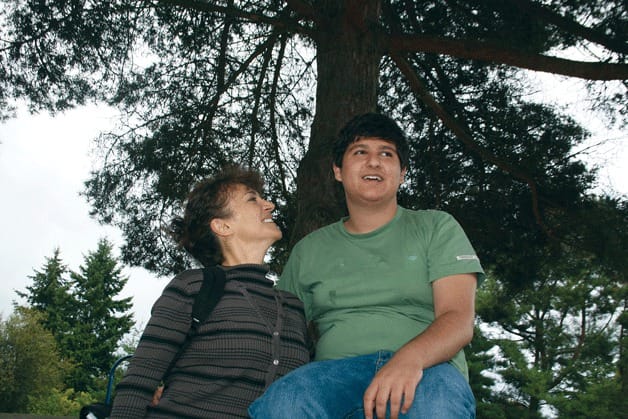 A woman and her adult son in front of a tree. The son is sitting up high, and the mom is looking up at him. Both are smiling.