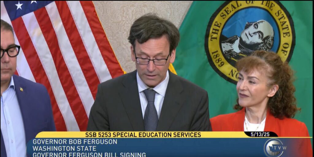 A photo of Governor Bob Ferguson reading a bill, with Arzu Forough standing next to him smiling. The American flag and Washington State flag are behind them.